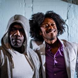 Two black men look directly at the camera. In the foreground, Benny stands in a grey hoodie and white shirt. Gil, wears a grey suit jacket and purple dress shirt, smiles, and wraps his arm around Benny's shoulder..