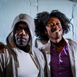 Two Black men are standing, one with his arm around the other, who has his hoddie up and looks off to the left with a serious face. The other, looks off right, smiling, in a grey suit with purple shirt.
