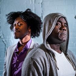 Two Black men stand one behind the other. In the foreground, Benny, played by Dwayne Alistair Thomas, looks directly at the camera with a more serious expression. Behind him, Gil, played by Garrick Vaughan, looks directly at the camera, his face more relaxed. Benny wears a grey hoodie. Gil wears a grey suit with a purple shirt.