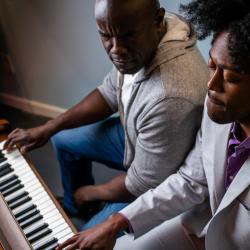 Two Black men sit at a piano. On the left, Benny's brow is furrowed. On the right, Gil looks down, but his face is peaceful.