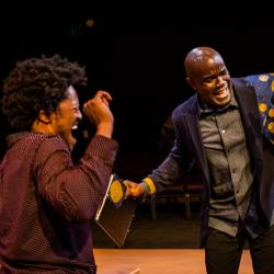 Two black men are smiling and dancing. On the left, Gil wears a burgundy dress shirt. On the right, Benny dances in a navy blazer with a blue and yellow wax print feature on the shoulder, worn over a grey dress shirt.