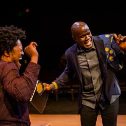 Two black men are smiling and dancing. On the left, Gil wears a burgundy dress shirt. On the right, Benny dances in a navy blazer with a blue and yellow wax print feature on the shoulder, worn over a grey dress shirt.
