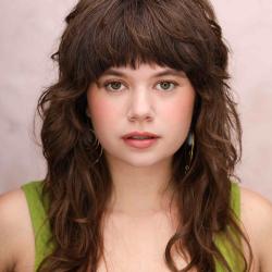 Anna Faye LIeberman's headshot. A young white person with wavy brown hair and bangs with brown eyes, wearing a green tank top and light makeup