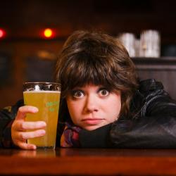 A young white woman rests her head on her arm on a bar, pint of beer in hand.