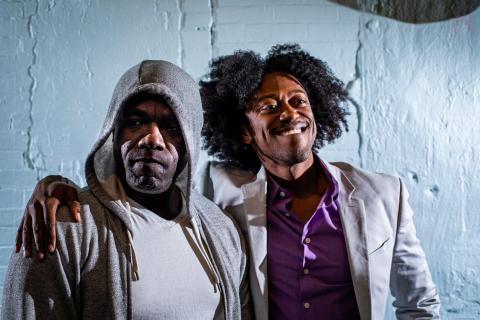 Two black men look directly at the camera. In the foreground, Benny stands in a grey hoodie and white shirt. Gil, wears a grey suit jacket and purple dress shirt, smiles, and wraps his arm around Benny's shoulder..